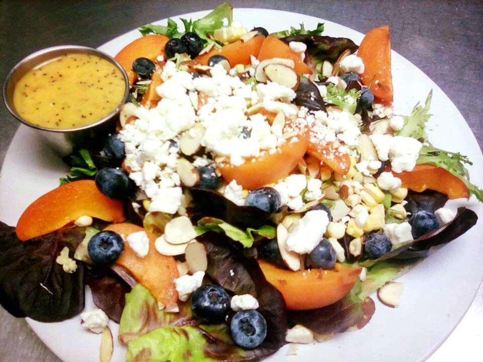 Fresh garden salad featuring mixed greens, sliced apricots, blueberries, almonds, and crumbled feta cheese, served with a side of tangy dressing.