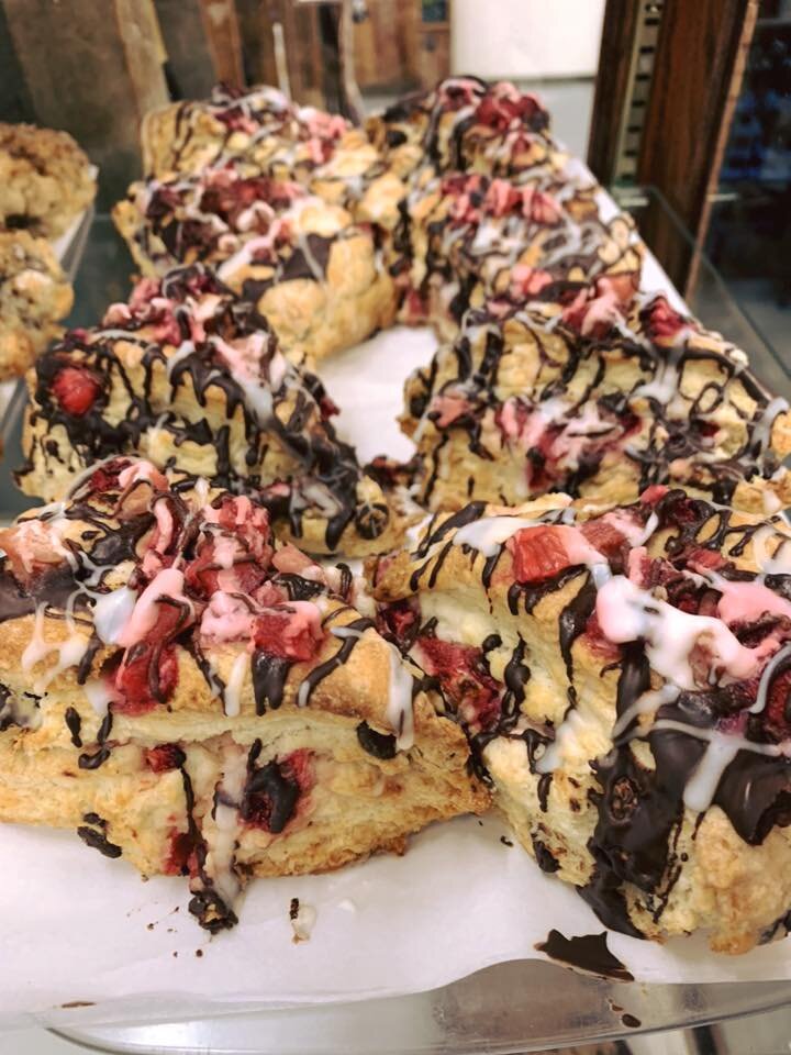 Freshly baked scones topped with chocolate drizzle, pink icing, and chunks of strawberries, displayed in a bakery case. Perfect for a sweet treat or dessert.