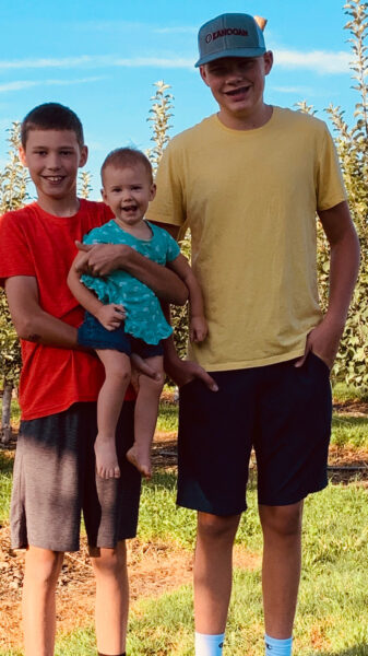 Three children stand together in an orchard, smiling and enjoying a sunny day. The two older boys wear casual t-shirts and shorts, while the younger girl is held by one of the boys, wearing a playful outfit. Lush green trees and a clear blue sky create a cheerful backdrop.