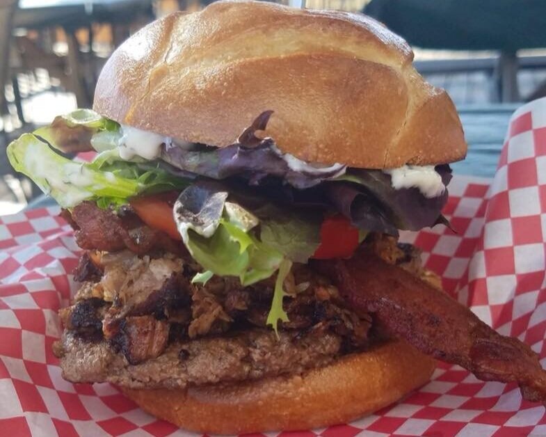 Juicy gourmet burger stacked with grilled meat, crispy bacon, fresh lettuce, tomato, and mayonnaise, served on a toasted brioche bun with red checkered paper background.