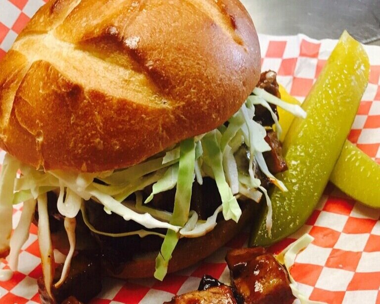 Close-up of a savory barbecue sandwich featuring tender meat, topped with fresh shredded lettuce, served on a golden brioche bun, accompanied by pickles and a side of diced barbecue meat on a red and white checkered paper. Perfect for food lovers seeking delicious comfort food.