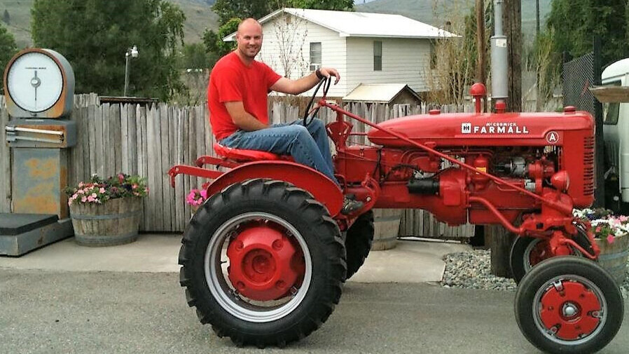 Man sitting on a vintage red Farmall tractor, wearing a red shirt and blue jeans, with a rustic wooden fence and flower pots in the background. A vintage scale is visible nearby, adding to the agricultural setting.